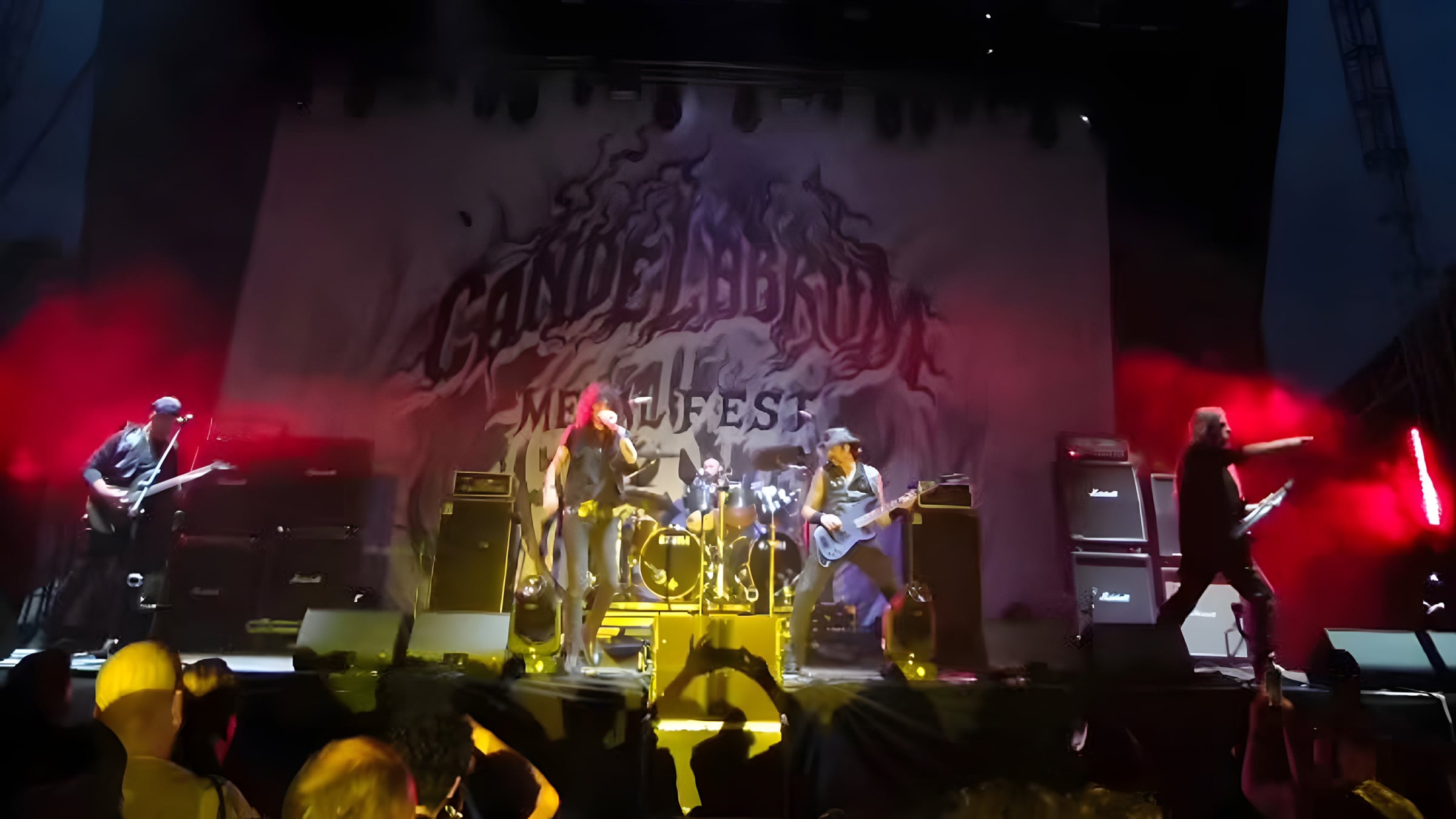 The whole band on the festival stage bathed in red light, with the crowd in the foreground and stacks of Marshall amps either side.