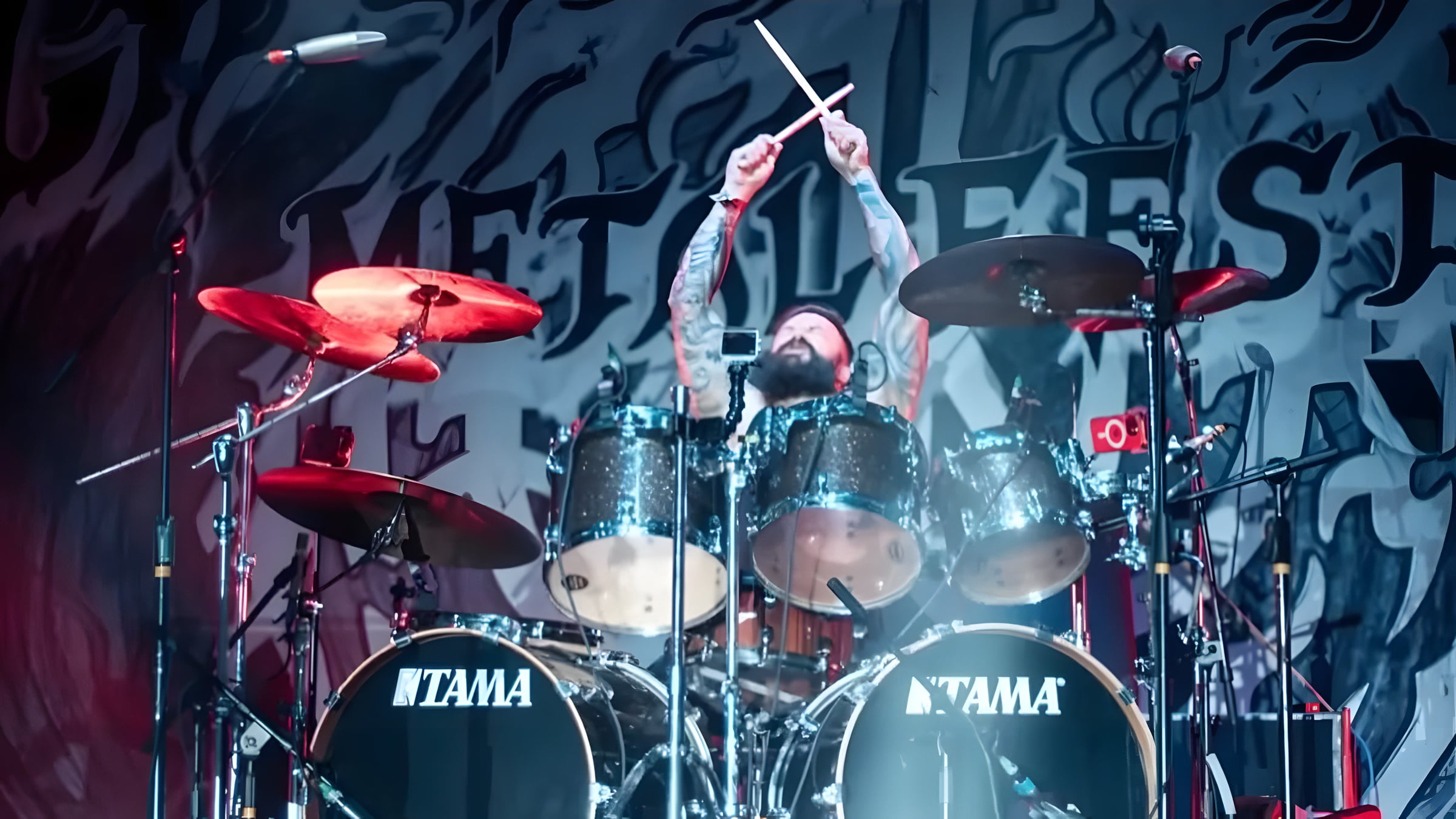 Logan Cade behind his drum kit with both sticks raised high, red-lit cymbals in front of the festival backdrop.